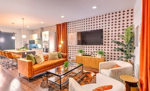 A modern living room with a sofa, coffee table, and chairs, a kitchen in the background, and a TV mounted on the wall.