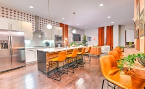 Una cocina moderna con electrodomésticos de acero inoxidable, una isla central con taburetes y un comedor con sillas color naranja.