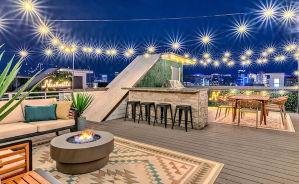 A rooftop bar with a fire pit, seating, and a city skyline view.