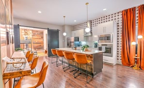 A modern kitchen with a central island, stainless steel appliances, and a dining area with orange chairs.