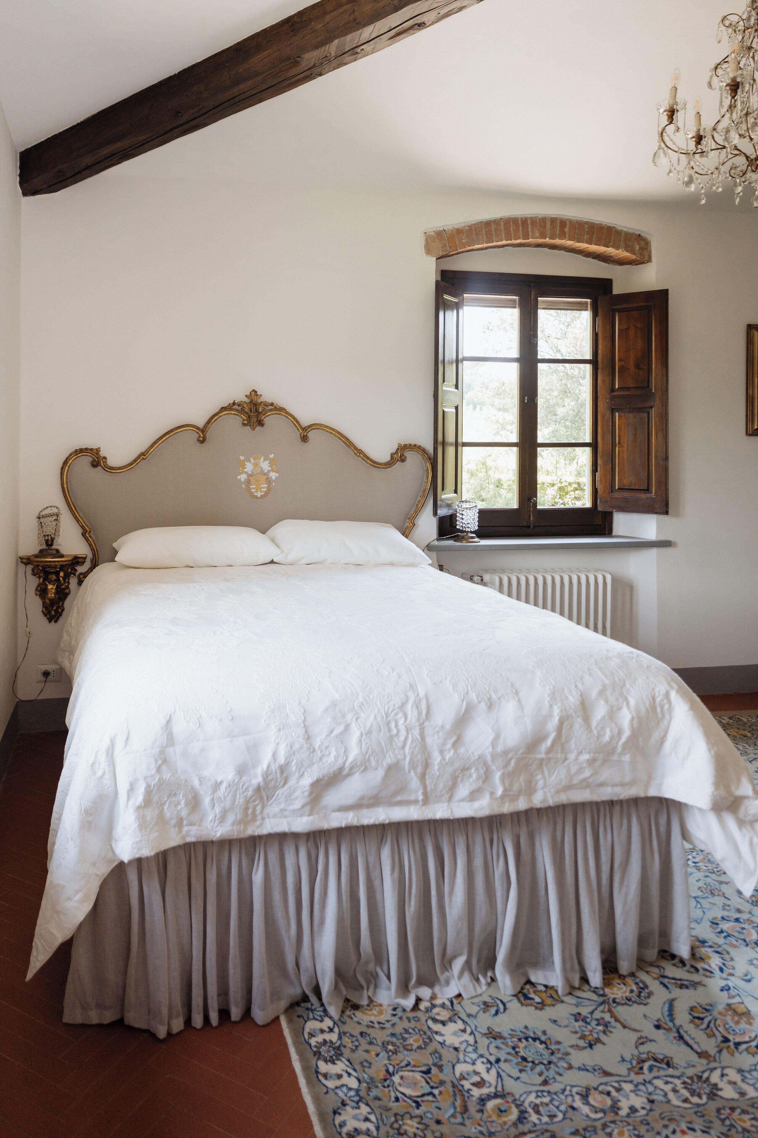 Une chambre à coucher avec un grand lit, une tête de lit en bois, une fenêtre avec des volets, un radiateur et un lustre.