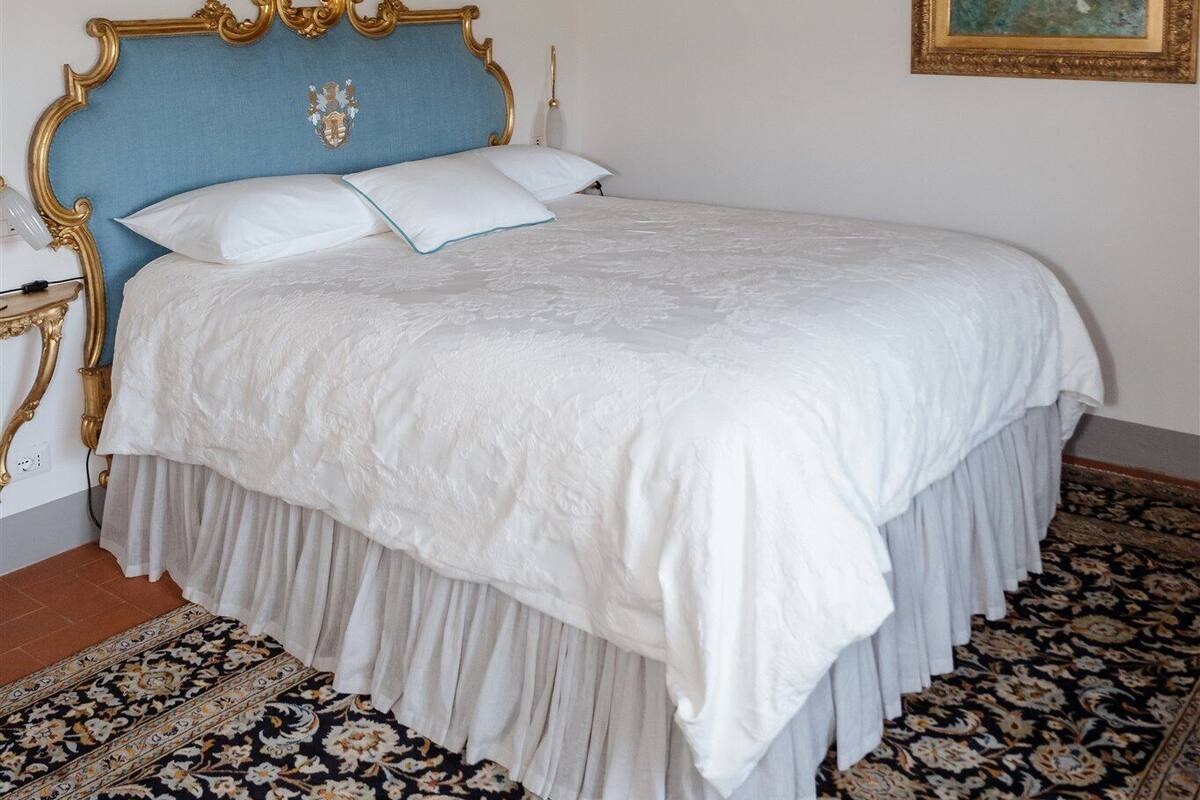 A bedroom with a large bed, ornate headboard, a chandelier, a painting, and a patterned carpet.