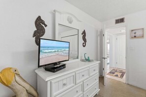 Una habitación con una televisión sobre un tocador blanco, una puerta que da a otra habitación y un adorno de pared decorativo.