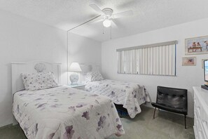 Ein Schlafzimmer mit zwei Betten, einem Deckenventilator, einem Fenster mit Jalousien und einem Sessel.