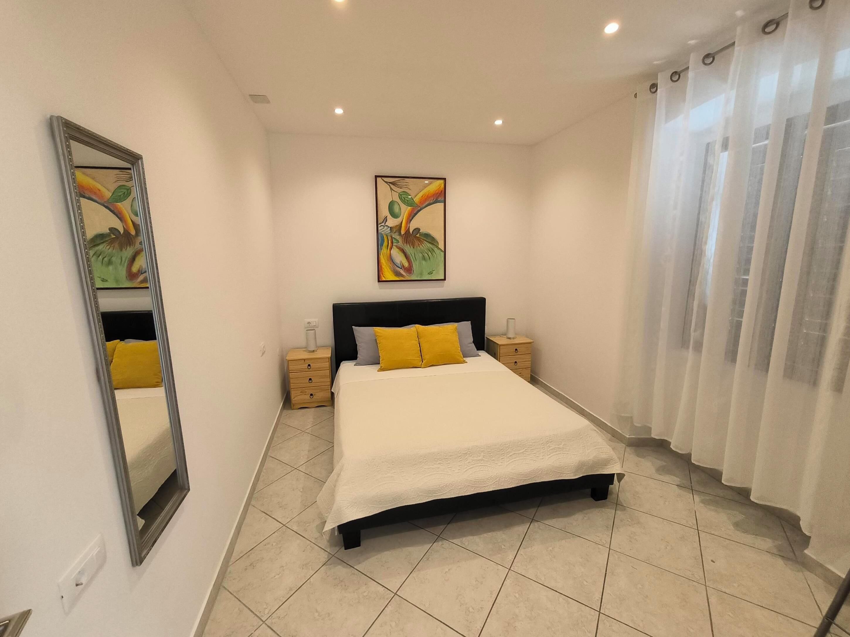 A bedroom with a bed, a nightstand, a mirror, a painting, and a window with curtains.