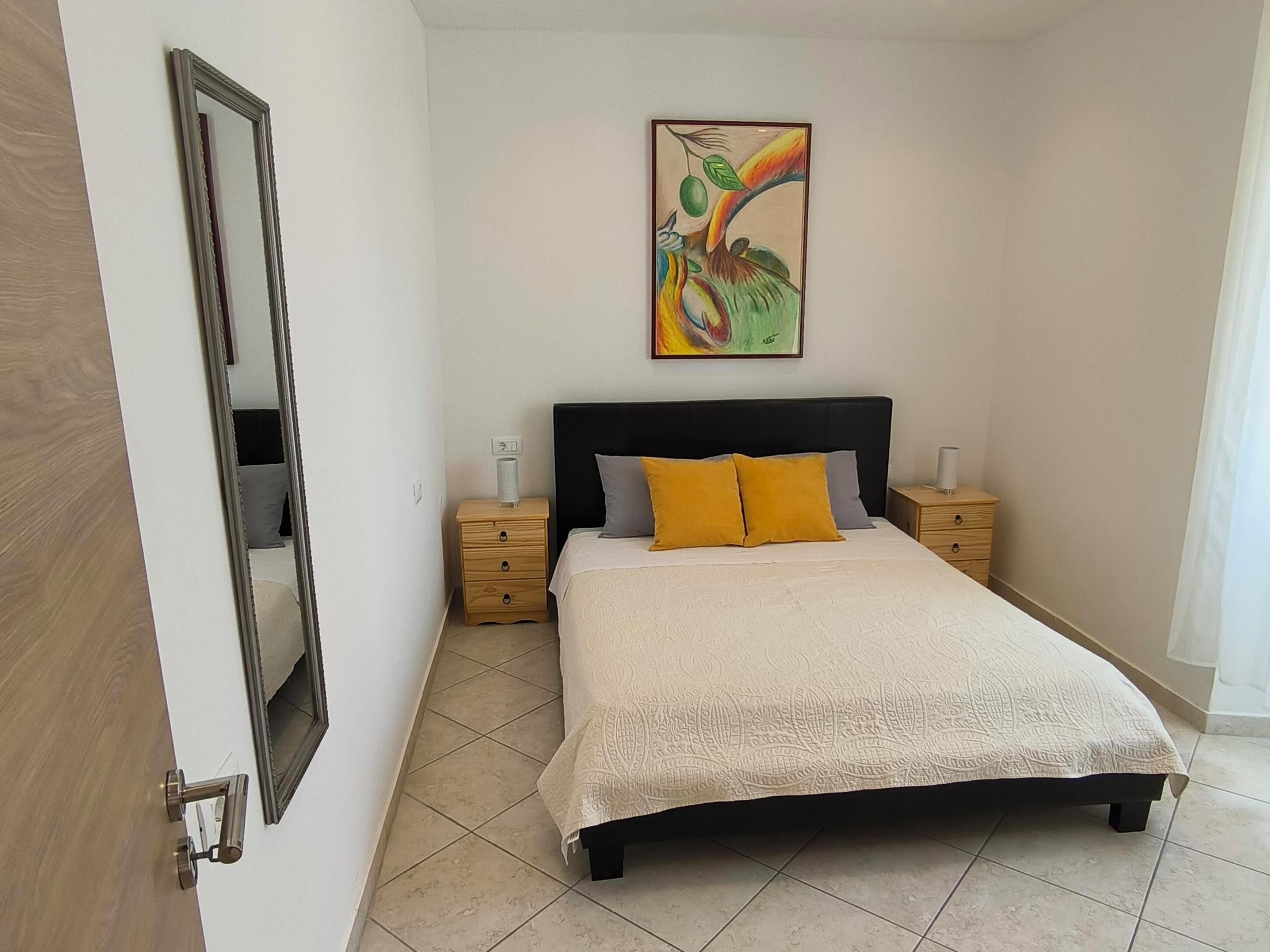 A bedroom with a bed, two bedside tables, a nightstand, a wall-mounted painting, and a door.