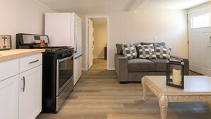 Eine kompakte Küche mit Herd, Backofen und weißen Schränken. Im Wohnbereich steht ein graues Sofa mit gemusterten Kissen, und ein kleiner Tisch mit Kerze ist zu sehen.