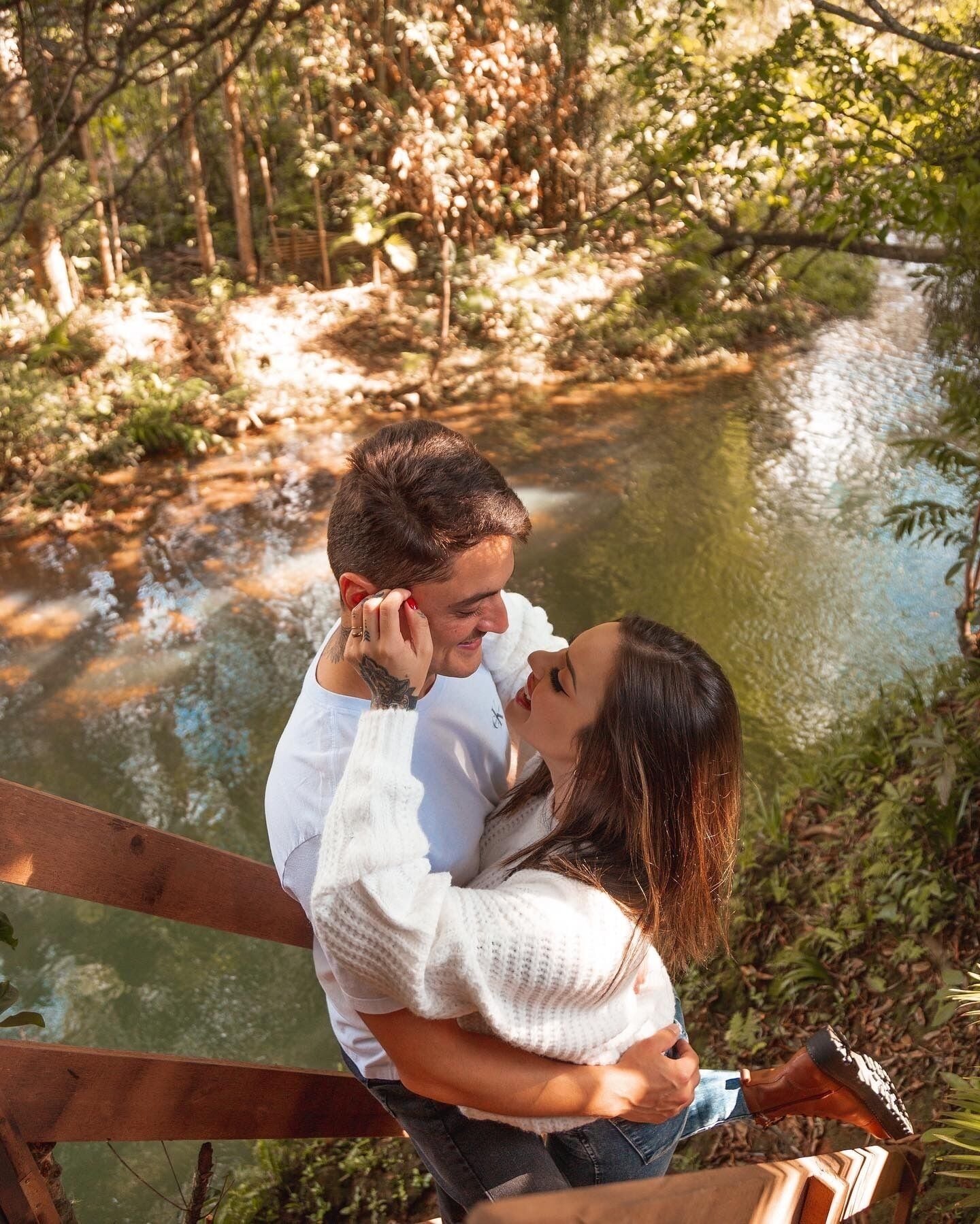 A couple embracing on a wooden bridge over a calm river, surrounded by lush greenery.
