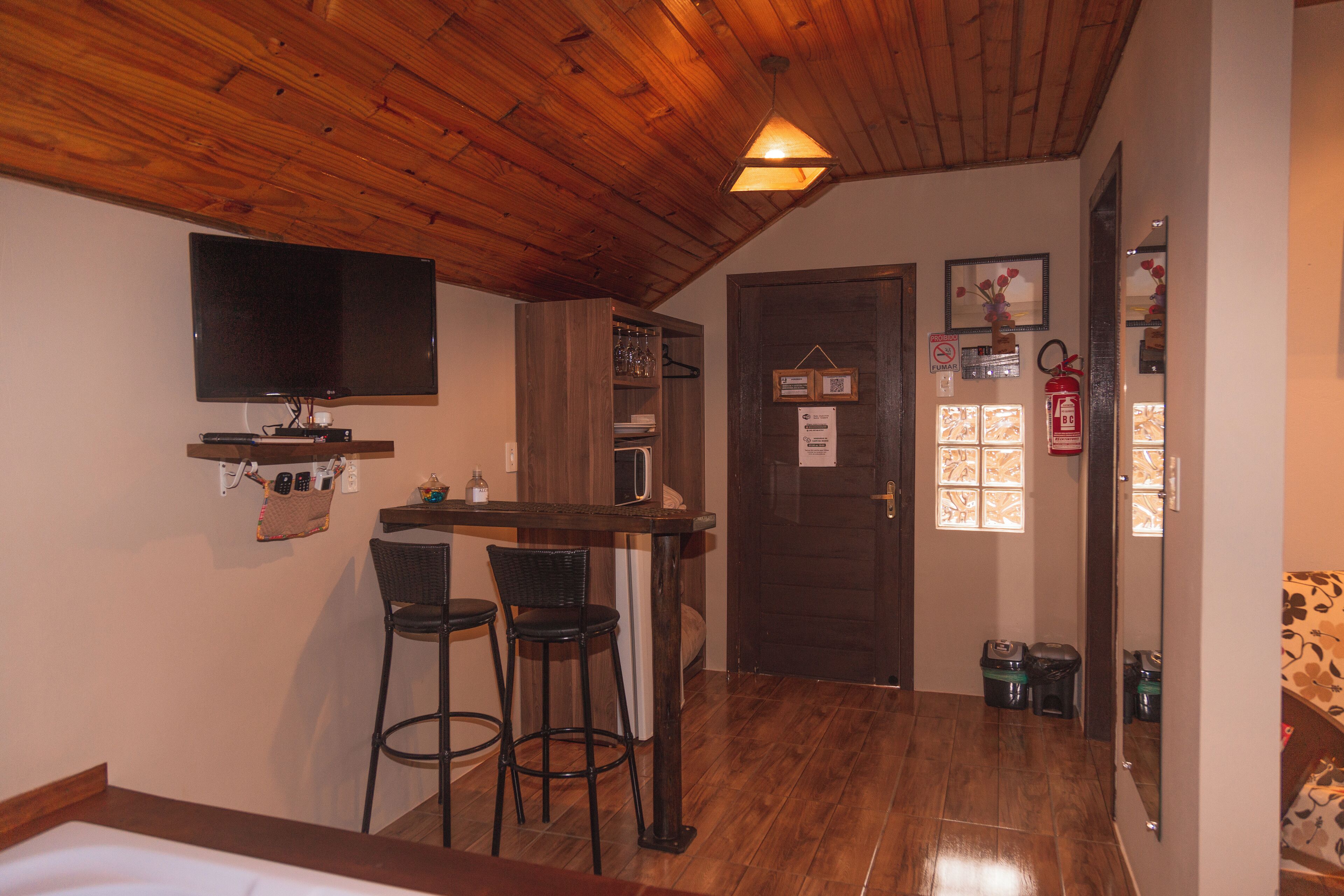 A room with a wooden ceiling, a TV mounted on the wall, a small bar area with stools, and a door with a fire extinguisher.