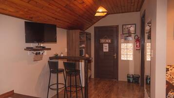 A room with a wooden ceiling, a TV mounted on the wall, a small bar area with stools, and a door with a fire extinguisher.