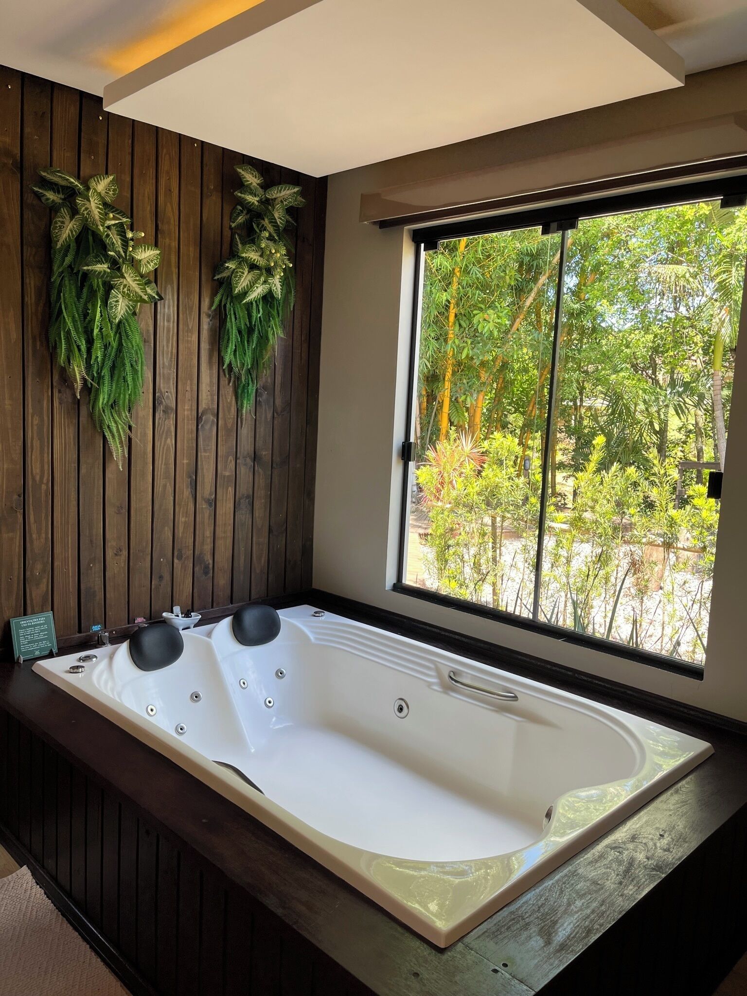 Un cuarto de spa con una bañera blanca grande, paneles de madera y vistas a un jardín exuberante a través de un amplio ventanal.