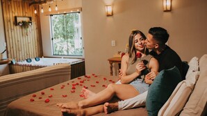 A couple sitting on a bed with rose petals, a wooden wall, and a window with a view of greenery. - Pousada Recanto dos Canyons (Praia Grande)
