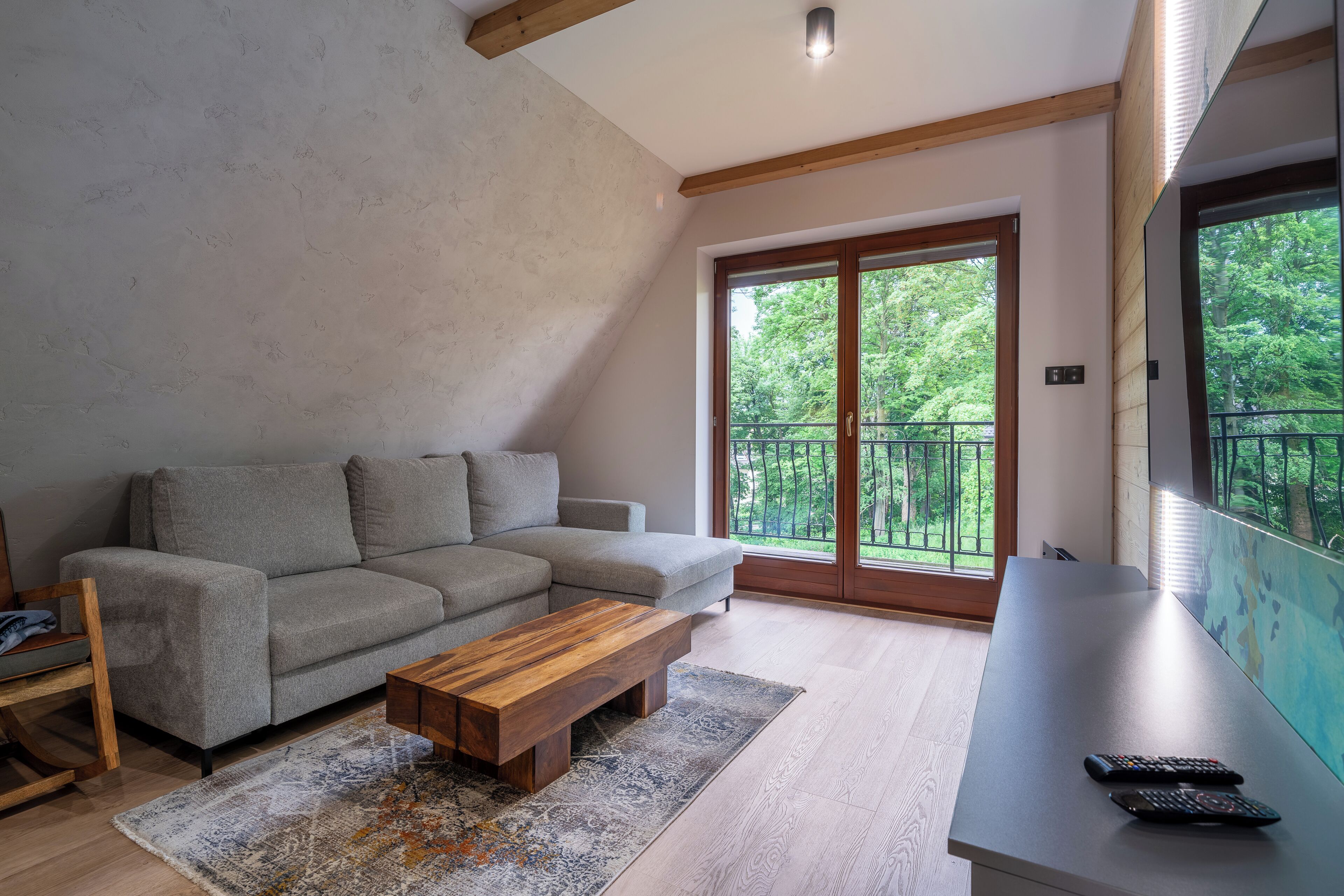 A modern living room with a grey sofa, a wooden coffee table, and a large window with a view of greenery.
