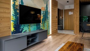 Un soggiorno moderno con una TV a schermo piatto montata su una parete decorata con un murale a tema naturalistico, un mobile TV grigio, un tavolino in legno e una porta in legno sullo sfondo.