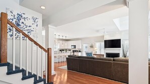 A modern living room with a wooden staircase, a grey sofa, and a kitchen in the background. - Spacious 4 Bedroom, Sleeps 10. Walk everywhere! (Bethany Beach)