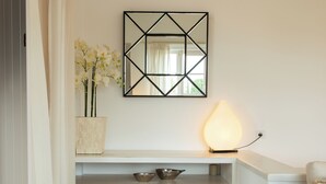 A minimalist living room with a white sofa, a geometric mirror, a lit table lamp, and a shelf with decorative items.