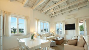 A spacious living area with a high ceiling, large windows, a flat-screen TV, and a dining table with chairs.