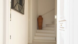 A hallway with white walls and a wooden floor, leading to a staircase with a large vase on the landing.