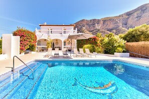 Una piscina con agua cristalina, rodeada de sillas de descanso y un edificio blanco con balcón.