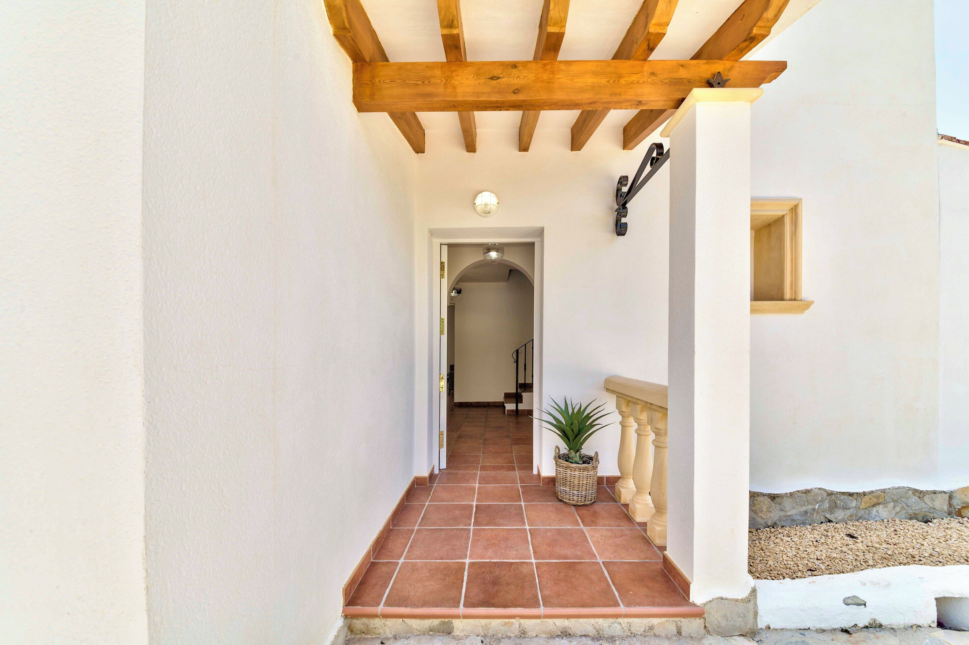 A corridor with a tiled floor leading to a doorway, a wooden ceiling beam, a wall-mounted light fixture, and a small potted plant.