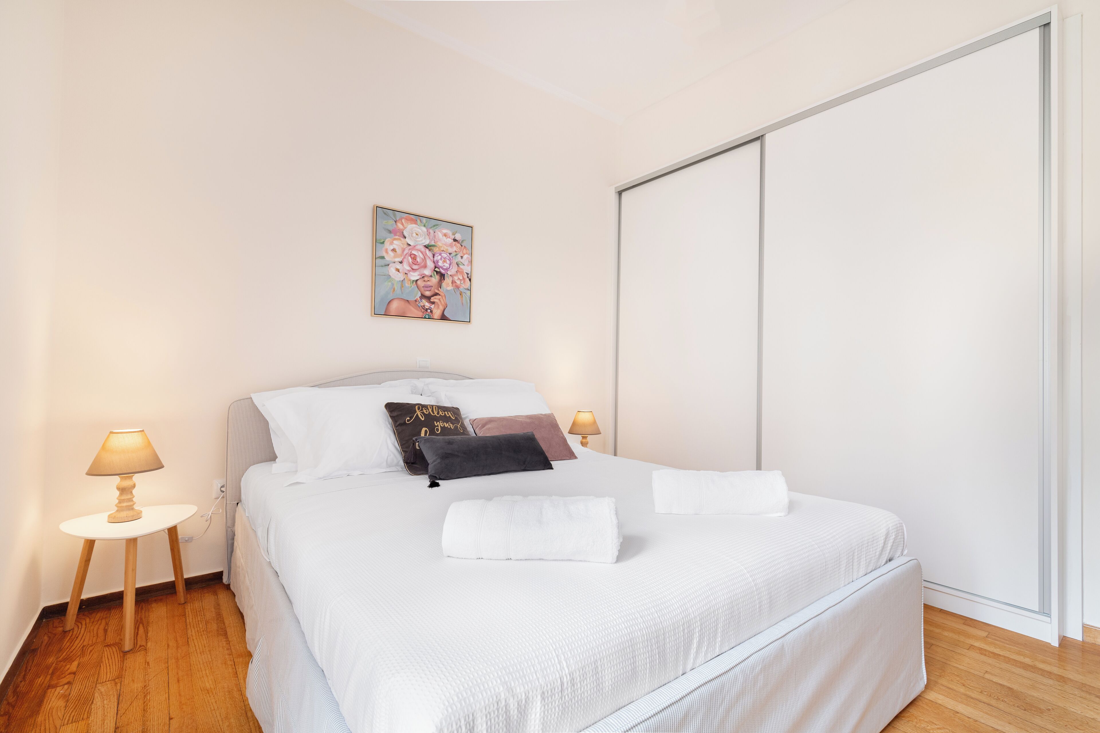 Een net opgemaakt bed met witte lakens, een decoratief kussen en een opgevouwen handdoek. Twee bedlampen staan op houten nachtkastjes. Een schilderij van bloemen hangt aan de muur boven het bed.