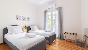 A hotel room with two beds, a wooden floor, a window with curtains, a wall-mounted air conditioner, and a small table with a plant. - Edem Beach Apartment by Alimos Marina (Alimos)