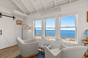 A room with a view of the sea, featuring a white door, a lamp, two chairs, and a small table.