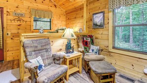 Ein gemütliches Blockhaus-Interieur mit Holzwänden, einem großen Fenster mit Blick auf einen Wald, einem gemusterten Sessel, einem kleinen Tisch mit Lampe und einer gesteppten Bank.