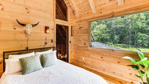 Ein Schlafzimmer einer Holzhütte mit einem großen Bett, einer Stierkopf-Dekoration und einem Fenster mit Blick auf einen Wald.