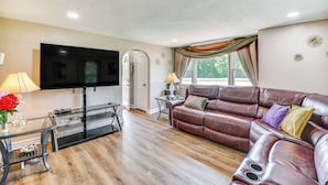 A living room with a leather sofa, a flat-screen TV, a glass coffee table, and a window with curtains.