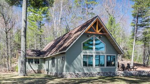 A cabin with a stone foundation, large windows, and a wooden roof structure, surrounded by trees. - 'northwood Tours Escape': Hot Tub & Lake Access! (Minocqua)