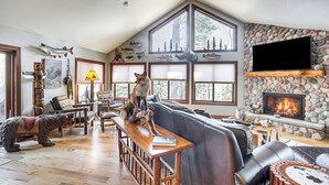 A spacious living room with a stone fireplace, large windows, and a dog on a bench. - 'northwood Tours Escape': Hot Tub & Lake Access! (Minocqua)