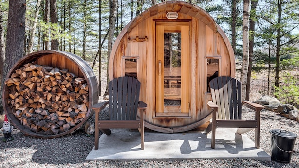 A wooden cabin with a door, two Adirondack chairs, and a barrel of firewood. - 'northwood Tours Escape': Hot Tub & Lake Access! (Minocqua)