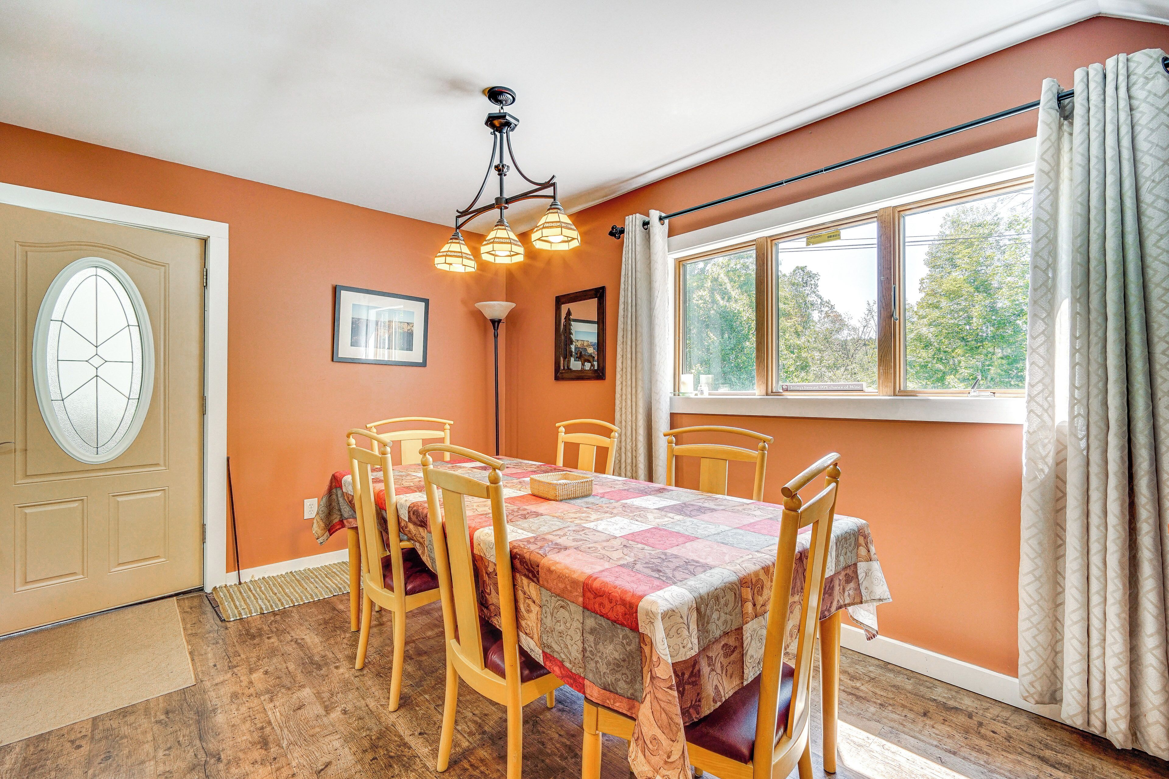Une salle à manger avec une table dressée pour six personnes, des chaises en bois, un lustre, une fenêtre avec des rideaux et une porte avec un paillasson.
