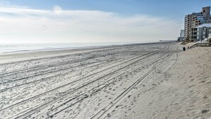 A beach with visible tire tracks and footprints, a clear sky, and a building with multiple floors and balconies on the right. - Walk to Ocean: Myrtle Beach Home w/ Screened Porch (Murrells Inlet)