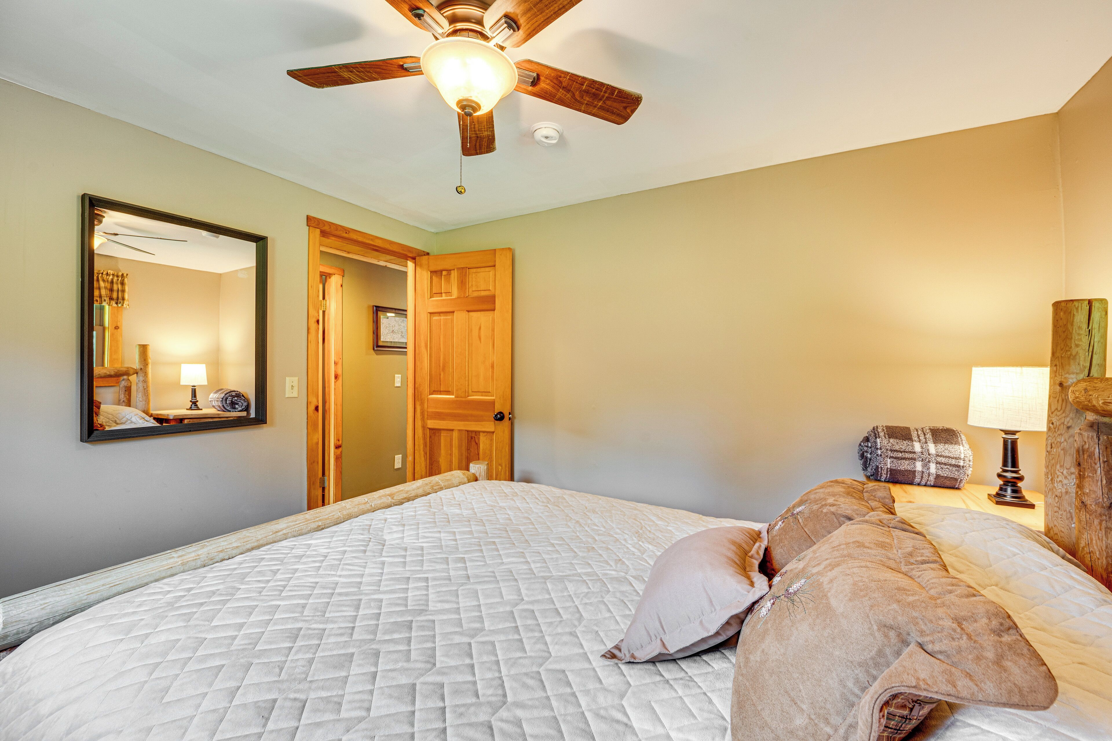 A bedroom with a bed, a wooden door, a ceiling fan, a mirror, and a nightstand with a lamp.