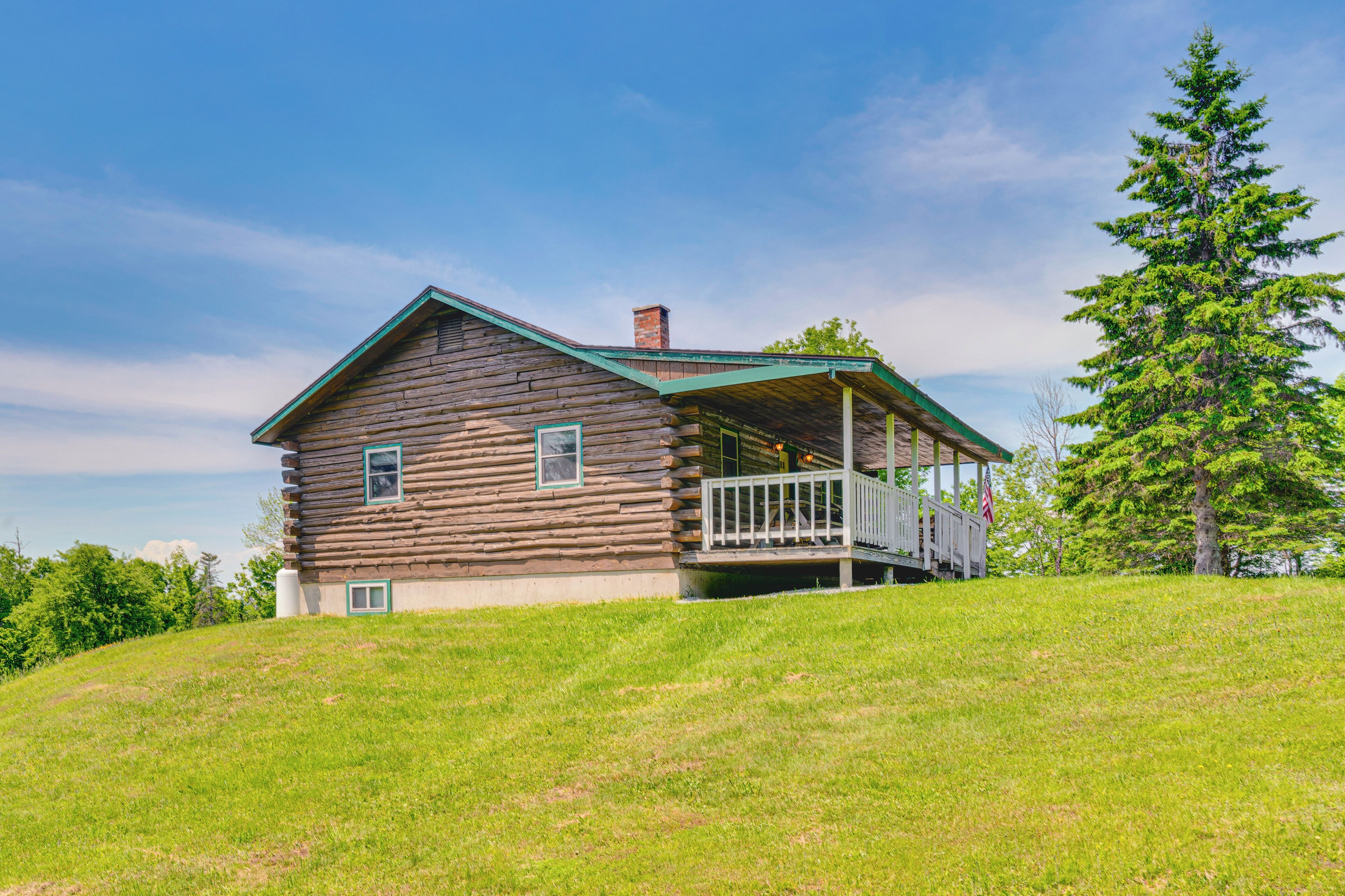 A log cabin with a porch, surrounded by a grassy hill and a tall evergreen tree.