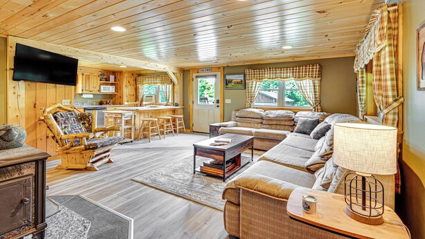 A cozy cabin interior with a wooden ceiling, a living area with a sofa and armchairs, a dining area with a table and chairs, and a kitchen with a microwave and a television. - Rustic Cabin in West Danville w/ Deck & Fire Pit! (West Danville)