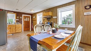 A dining area with a wooden table set for four, surrounded by white chairs. A kitchen is visible in the background with wooden cabinets and a window. - Charming Stone Cottage in Marion w/ Fire Pit! (Marion)