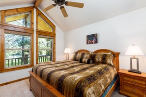 Un dormitorio con una cama grande, cabecera de madera, dos lámparas de noche, un ventilador de techo y ventanas amplias con vistas a árboles.