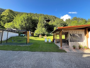 Un área de estar al aire libre con techo, que incluye una mesa, sillas y un columpio, con vistas a una colina boscosa.