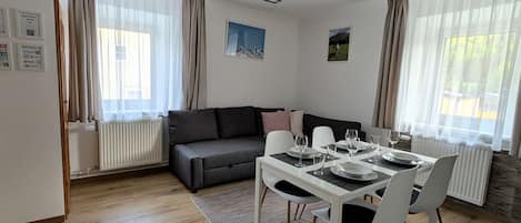 A modern living room with a dining area, a grey sofa, a white table set for four, and a large window with curtains.