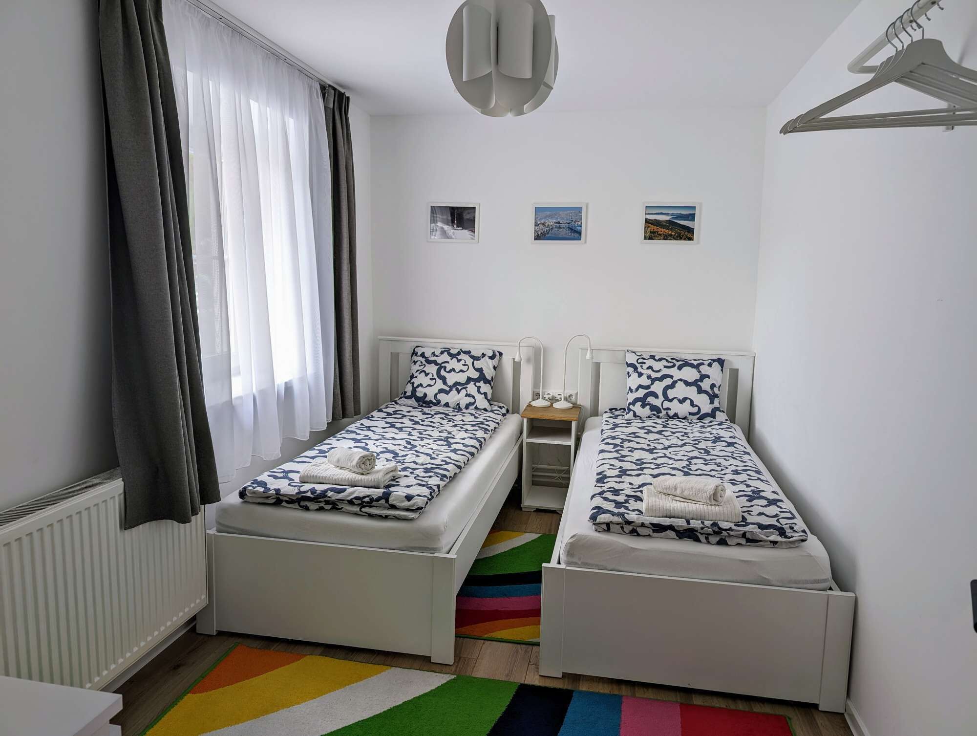 A twin room with two single beds, a radiator, a colorful rug, and a view of the city.
