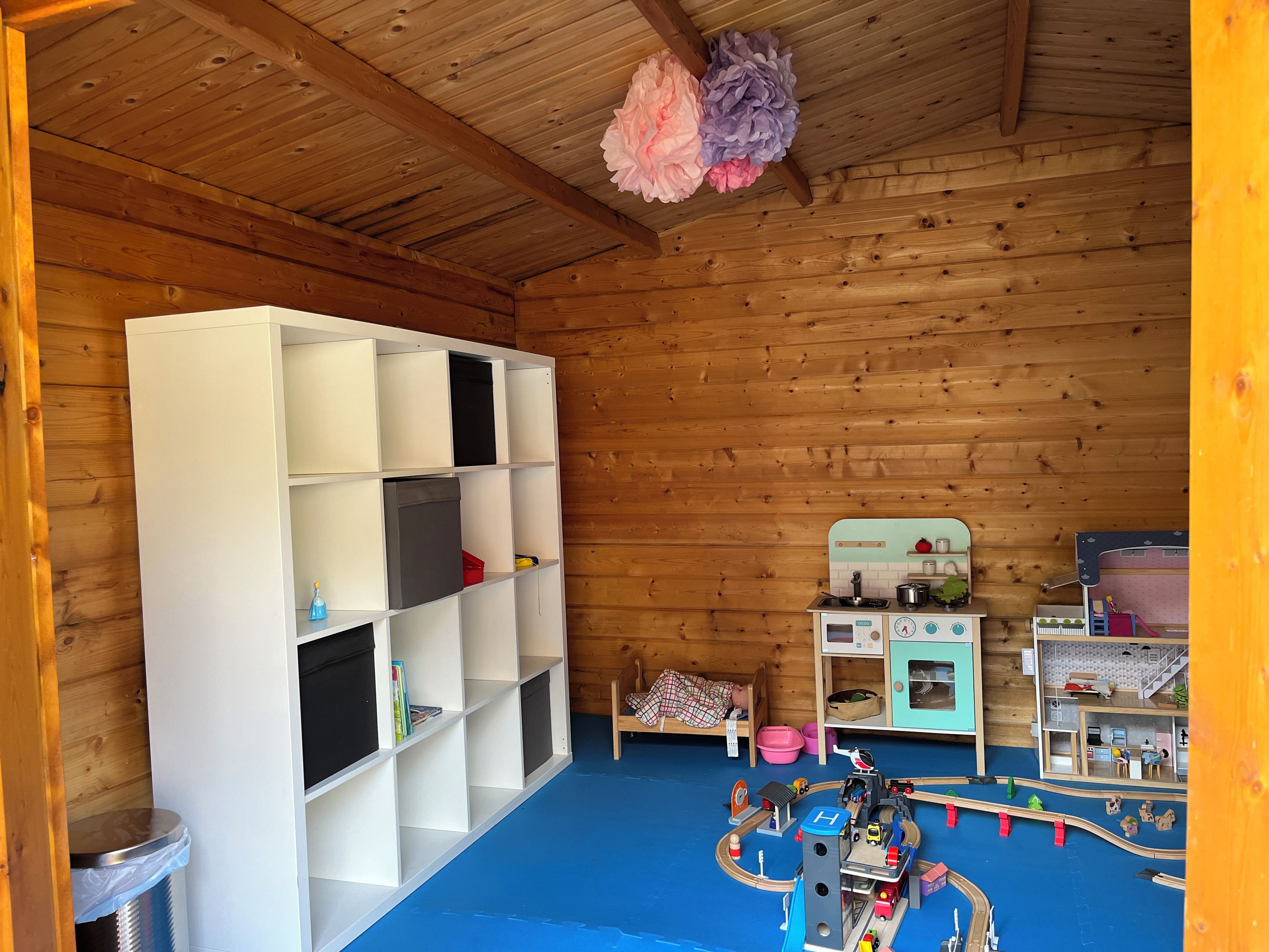 Een kamer met een houten plafond, een wit schapenkastje, een treinset en een kleine kitchenette.