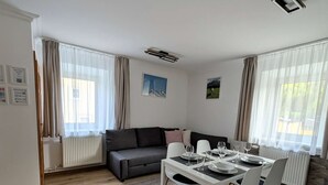 A modern living room with a dining area, a grey sofa, a white table set for four, and a large window with curtains. - UPSTAIRS, 2 BEDROOMS APARTMENT - small room with double bed (Bad Bleiberg)