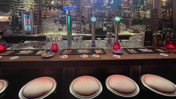 Um bar com vários bancos, uma variedade de garrafas e copos, e uma tela de televisão ao fundo.
