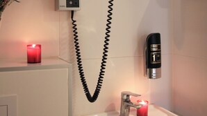 Casa de banho com secador de cabelo, dispensador de ar quente para as mãos, espelho, lavatório e uma vela vermelha.