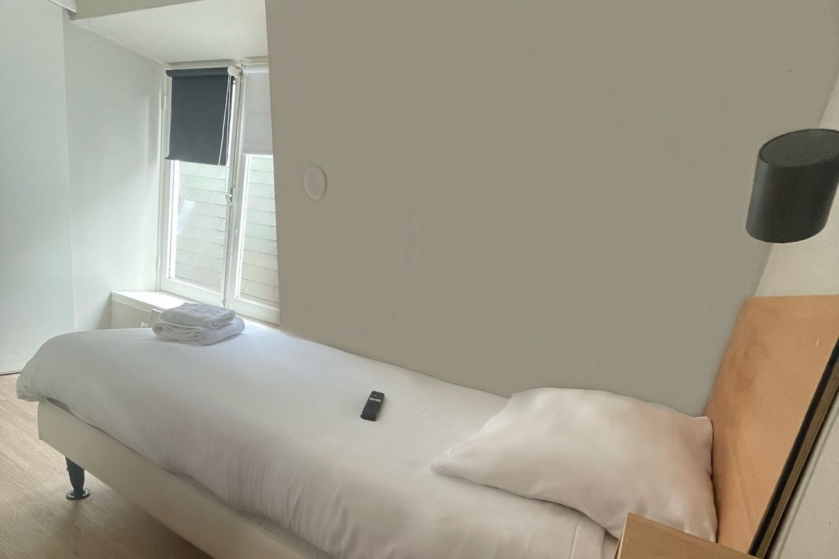 En enkeltseng med hvidt sengetøj, en fjernbetjening på sengen og et vindue med persienner.