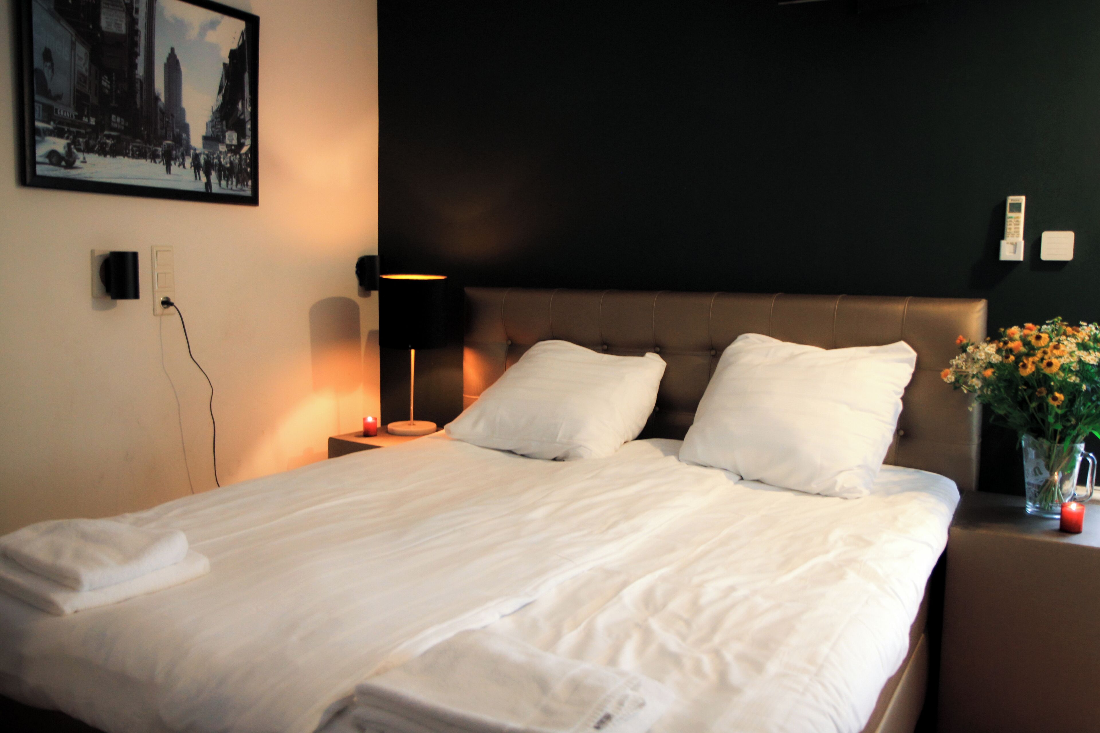 A neatly made bed with white linens, a dark headboard, a bedside lamp, and a wall-mounted picture.