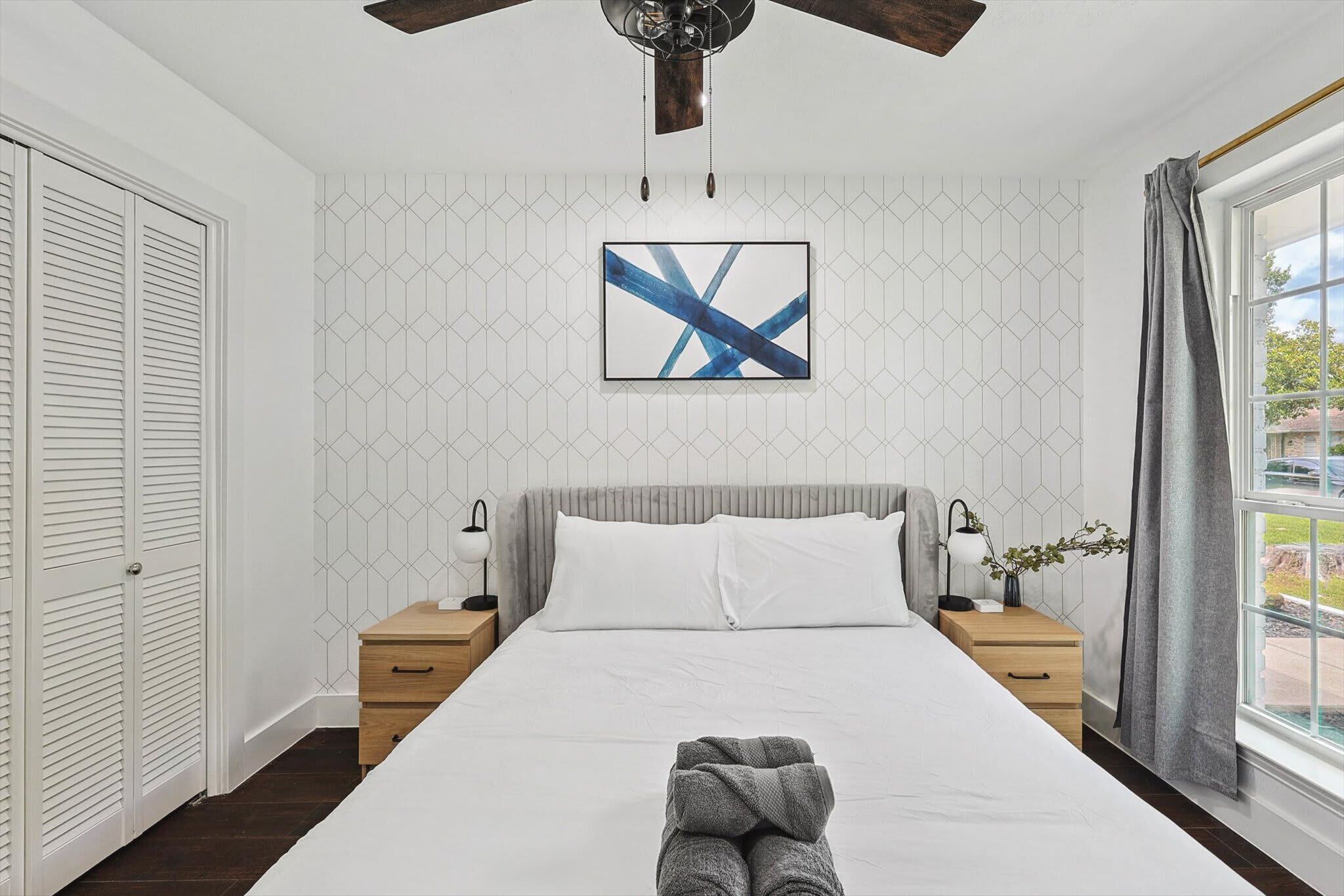 Un dormitorio ordenado con cama, mesitas de noche, ventilador de techo y ventana con cortinas.